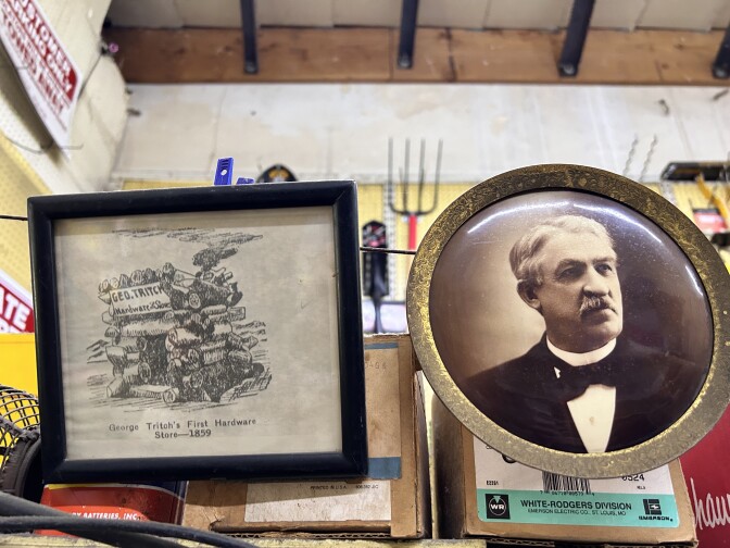 A close-up of a pair of framed photos. On the left, a black and white drawn depiction of a small wood cabin, with the words "George Tritch's First Hardware Store - 1859" printed below, in a simple square black frame. On the right, an aged gold circle frame surrounds a sepia-toned portrait of a man. The man is wearing a suit, with a bowtie, and has a neat graying mustache. 