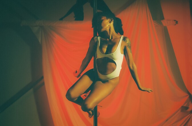 A feminine presenting person with medium-dark skin tone wears black curly hair to the side and a one piece white leotard with the belly cut out while balancing on a pole with their arms to the side. Behind them is a red curtain. 