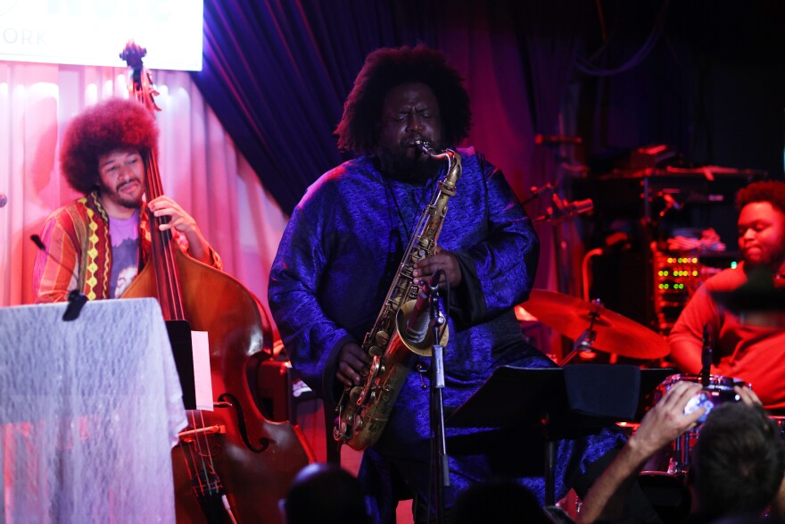 A Black man in an indigo colored outfit plays the saxophone into a microphone as other band members play their instruments behind him.