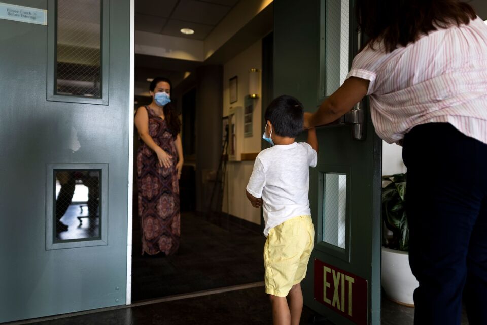 A young boy is shown in a doorway between a caregiver and his mother as she drops him off. The boy wears yellow shorts and a white t-shirt and holds the hand of the caregiver. His back is to the camera. 