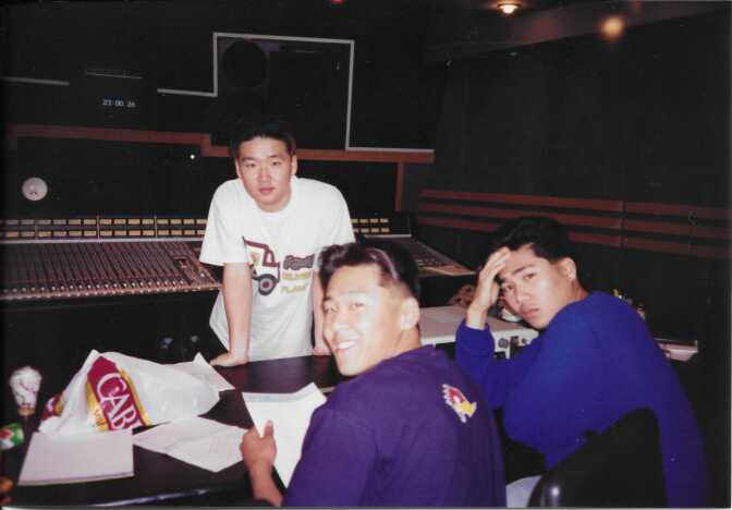 Three young Asian men with lighter skin tone work in a recording studio. One in a white shirt is standing and facing the camera, while two others are seated, back to the camera but looking over their left shoulders. One in the middle is smiling.