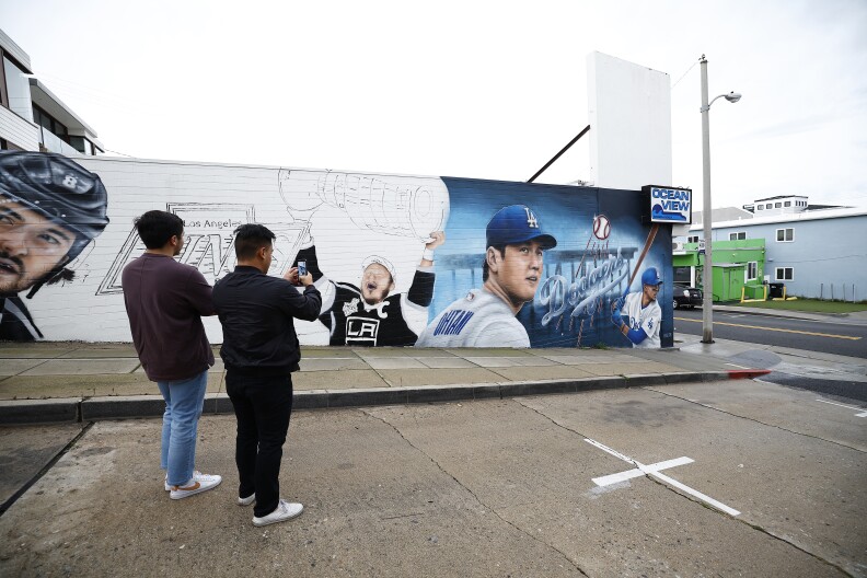 Two men with their backs to the camera take a photo of mural of Shohei Ohtani of the Los Angeles Dodgers, painted on the side of a liquor store.