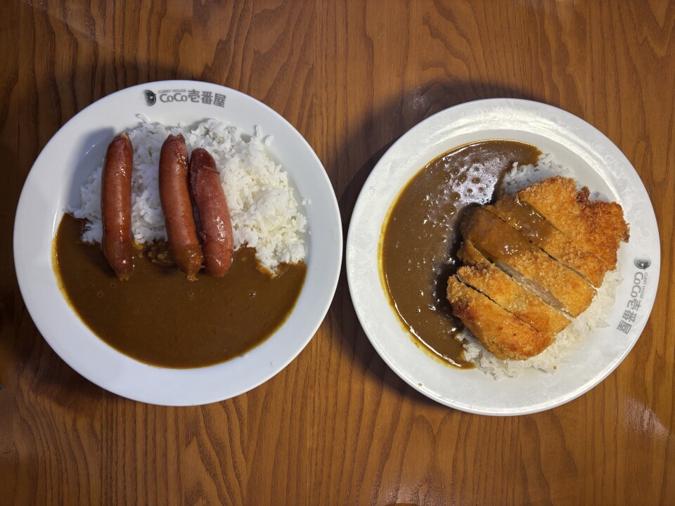 Two white plates sit against a brown wooden surface. The plate on the left contains a brown sauce with white rice and three cooked sausages on top. Next to it, the plate on the right contains the same brown sauce and white rice, with a light brown fried cutlet on top.  
