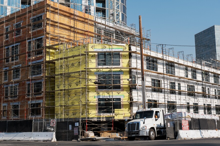 A new construction building, white, yellow, and wood-paneled, is covered in scaffolding during the daytime. A large truck sits in front of the structure. On section of the building is 3 stories, the other is 5 stories.