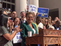 Mayor-elect Karen Bass, dressed in a green pantsuit, gives remarks at a wooden podium as male and female supporters of different races applaud. 