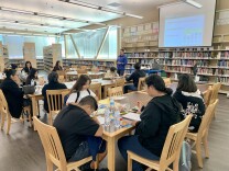 Adults and children sit at blond-wood tables in a large room surrounded by books as a man in the distance, standing, lectures beneath a projection screen.