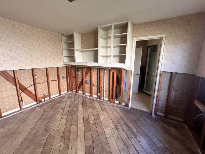 An interior room with wooden floors and white shelving affixed to one of the walls. The lower half off the walls are missing, exposing the lining of the home or the adjacent room.