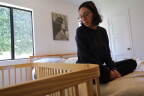 A woman with medium-tone skin and glasses looks down on a wood crib while sitting on a bed. The photo of a woman hangs on a white wall between windows.