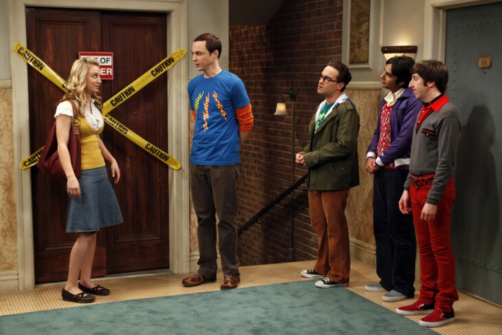 Four men speak to a woman with blonde hair in a skirt in a hallway where yellow caution tape blocks and out of order elevator.