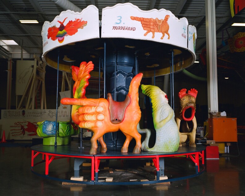 A mostly-assembled carousel features several fantastical and colorful seats, including a hand making a finger gun shape that has hooves attached.