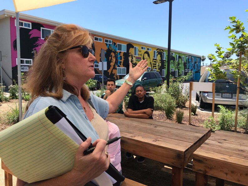 A woman holding a notepad speaks while waving her hand. There are people sitting at a table behind her.