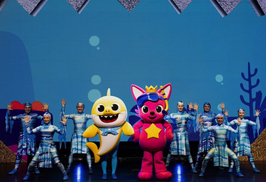 dancers surround two performers in baby shark and cat costumes on a stage.