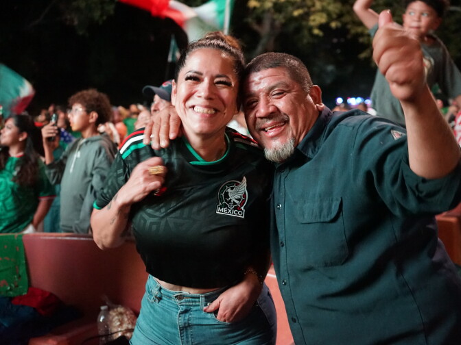 A woman and man stand arm in arm, grinning. Behind them, a crowd watches a large outdoor screen.