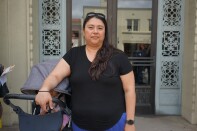 A woman with a medium skin tone wearing a black shirt and blue pants stands in front of a glass door with ornate metal lining on either side. She rests her right arm on a gray stroller. A baby's feet and torso can be seen under the stroller. The woman wears black sunglasses on her head and has long dark brown hair.