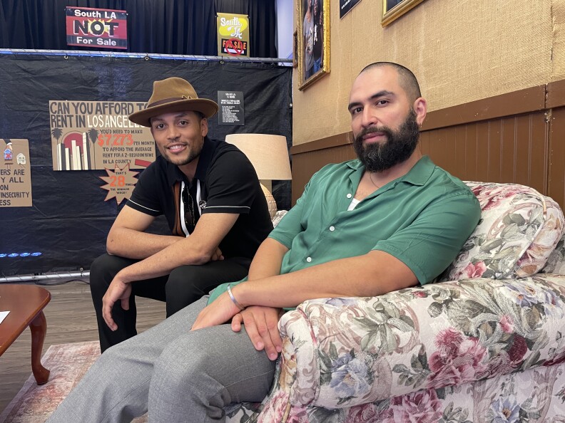 Two men, one with a hat, both with brown skin, sit on a couch in a space that's meant to look like a real living room.