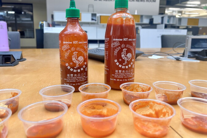 A smaller, mostly empty bottle of Sriracha and a larger, mostly full bottle sit behind two rows of clear used condiment cups. 