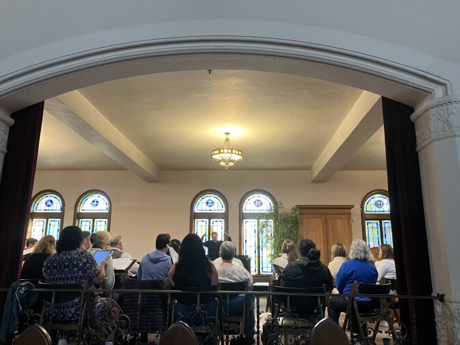 A light-skinned man stands in front of a group, hands raised above his head. He stands in front of two stained windows, with an indoor plant. Rows of people sit in chairs looking at him.