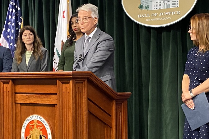  L.A. County DA George Gascón announces criminal charges standing at a wood podium. He wears a grey suit. 