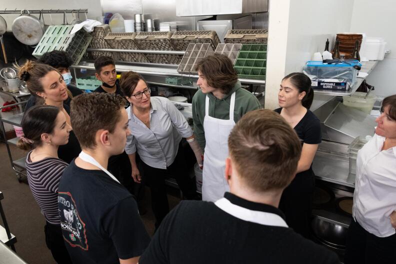 a woman in a white shirt talks to a group of 10 employees who all look to be in their 20s or teens, in a restaurant kitchen