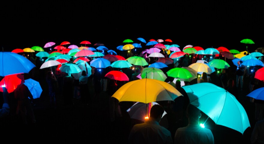 100 or so people hold bright, mono-colored umbrellas in a dark environment