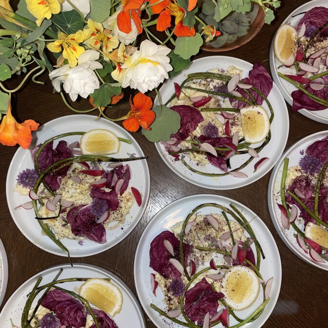 An overhead image of two rows of white ceramic plates each containing slices of radishes and beets on hummus., with lemon segments.  Next to the dishes are white, yellow, and bright orange wildflowers. 