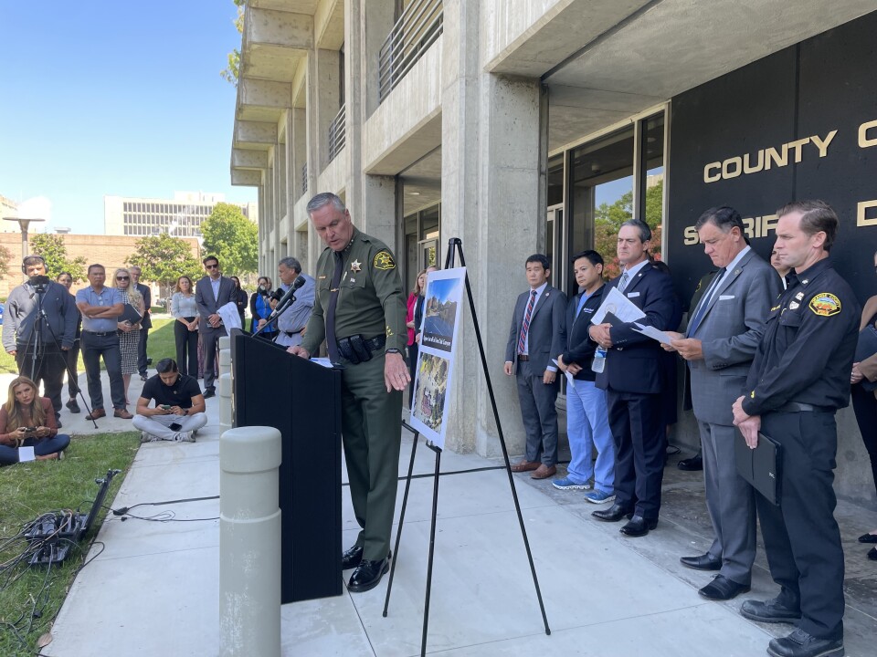 The sheriff in full uniform points at a set of photographs while describing the crime scene.