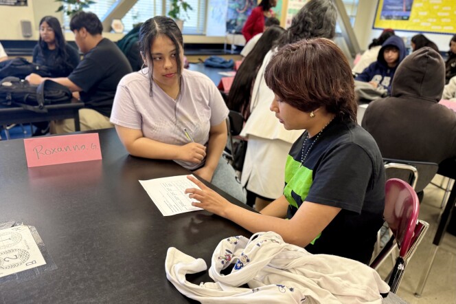 A young person with medium skin tone sits at a black table looking over a piece of paper. Next to them, a young woman with medium skin tone watches, with a sign next to her that says "Roxanna."