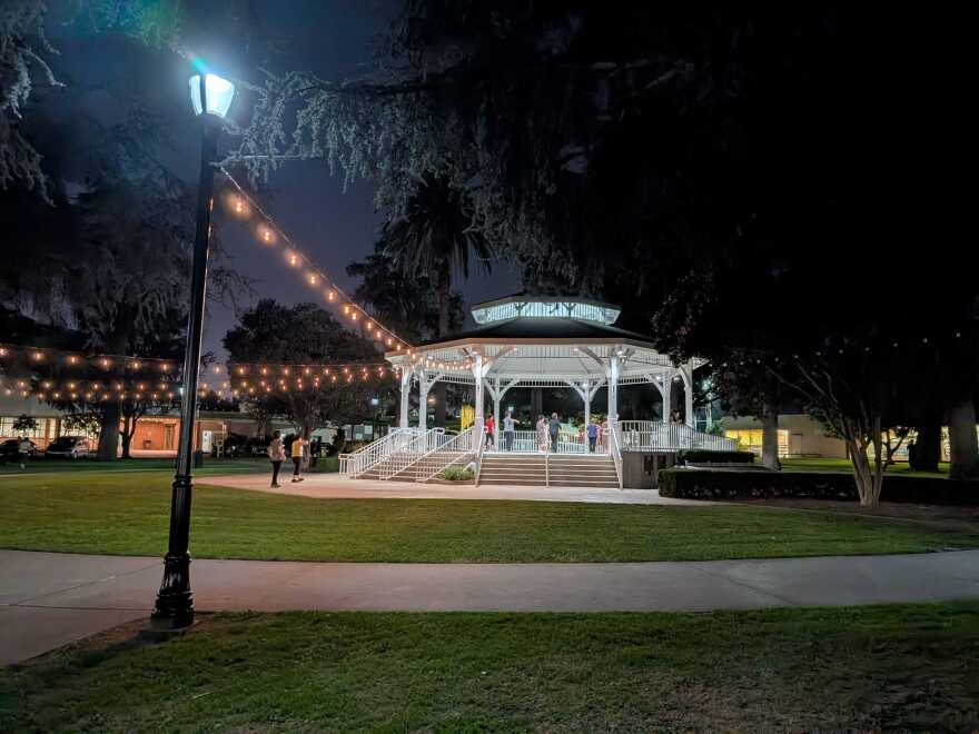 People inside a large gazebo in a park at night. The gazebo lights are on, as are strings of light between the gazebo and light poles.