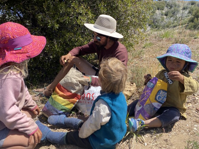 One little girl and two boys sit on the ground and watch their Dad pull food out of a rainbow-colored bag. One of the boys has a bag of pretzels in front of him. 
