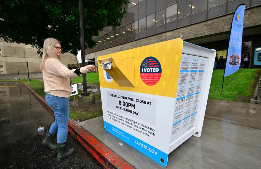 A middle-aged woman drops a ballot off outside a large yellow ballot box on a rainy Los Angeles street.