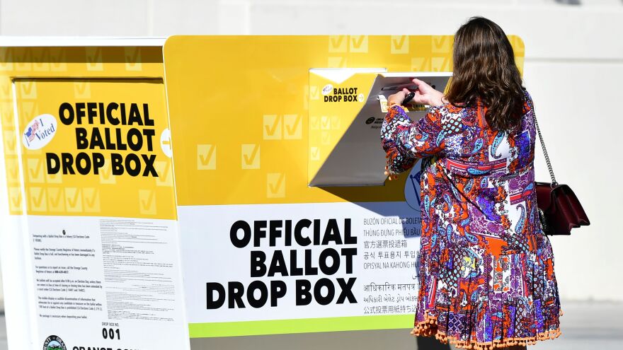 A person puts her ballot through the slot of an official drop box. The ballot box is orange and white with large black lettering.