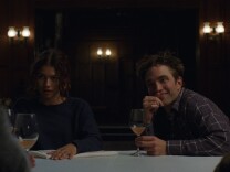 A female presenting person and a male presenting person sitting side by side at a table, across from two other figures who are not visible. Wine glasses are on the table.