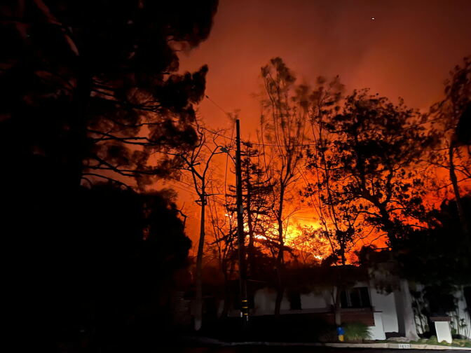 The yellow, orange, and red glow from a fire behind a home and trees at night 