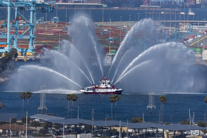 A fireboat sprays multiple arcs of water into the air from its cannons while cruising through a harbor. The background features an industrial port with cranes, containers, and various buildings. Palm trees and other structures are visible in the foreground along the waterfront.