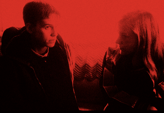 In a scene from the 1999 film “Boys Don’t Cry,” on the left side of the image, actor Hilary Swank plays trans man Brandon Teena. Her brown hair is cut short, and there is a serious expression on her face as she sits on a couch and looks at someone off camera. She is wearing a dark shirt with the collar of a white undershirt showing, and a puffy brown jacket on top. Next to Swank on the couch (covered in a multi-colored, chevron-pattern blanket) actor Chloe Sevigny (playing Brandon’s girlfriend, Lana Tisdel) is seen in profile, looking at Swank. She has long-ish reddish blonde hair and is wearing a black jacket with gray stripes down the arms.