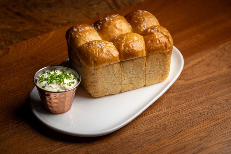 A white rectangular plate contains a small silver container of butter with herbs on top, and an old style loaf of bread made up of six rolls