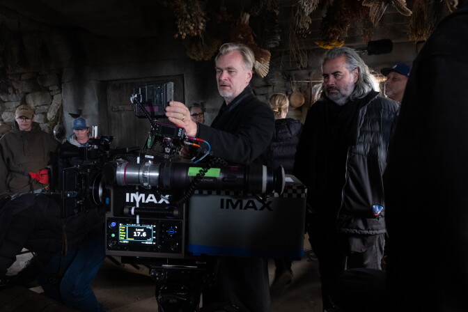 Director Christopher Nolan stands next to Cinematographer Hoyte van Hoytema on a dark film set, both looking into an enormous IMAX camera. Crew can be seen in the background.