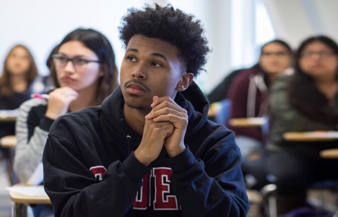 A male college student with medium skin tone wearing a dark gray hoodie sits at a desk in a classroom, with other students seated at desks in the background.