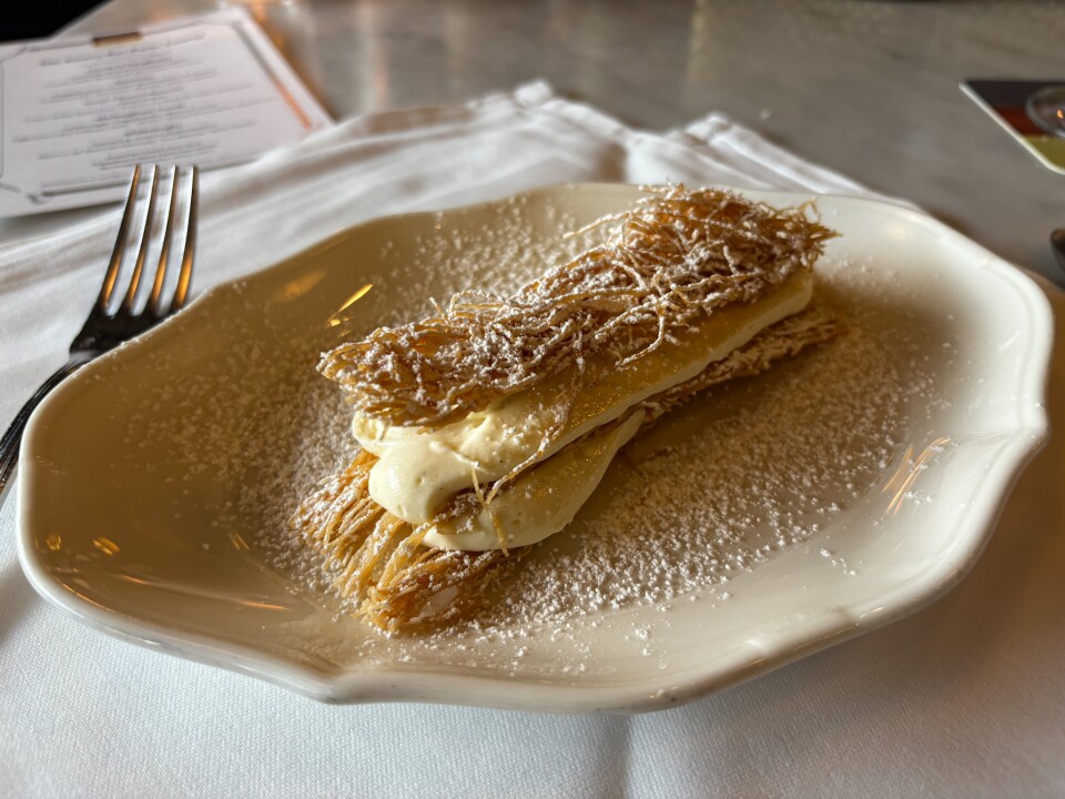 A refined mille-feuille containing multiple layers of ultra-crisp, golden pastry made from kataifi (shredded filo dough), with a large amount of thick white cream in between as its filling, and dusted with a heavy amount of white powdered sugar. 