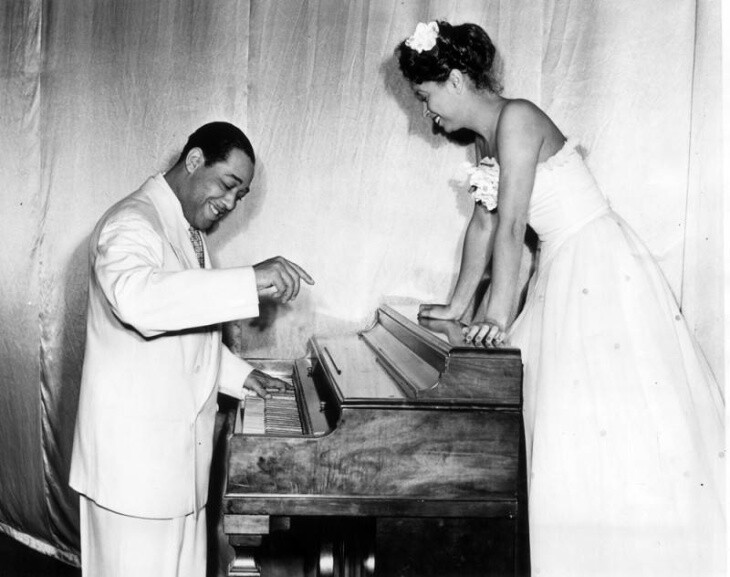 A Black man and woman face across from each other at a piano. Both are wearing white