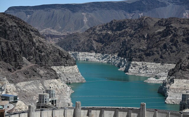 The rocky banks of a river show a white band toward the bottom, indicating a previously higher water level. The Hoover Dam is shown in the foreground.