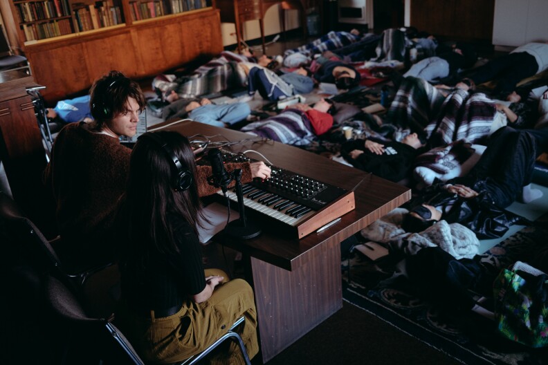 Collin Davis and Brittany Bueno lead a music listening session at the Philosophical Research Society in Los Feliz. They sit at a table with headphones on, adjusting knobs on a synthesizer. A dozen or more people lay on the floor listening. 