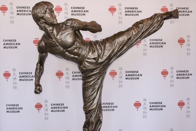 A fiberglass statue of martial arts legend Bruce Lee is set against a backdrop printed with a red lantern logo for the Chinese American Museum.