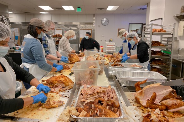 Eight people stand around a large table carving many cooked turkeys in a commercial kitchen. They wear protective masks, hairnets, and aprons.