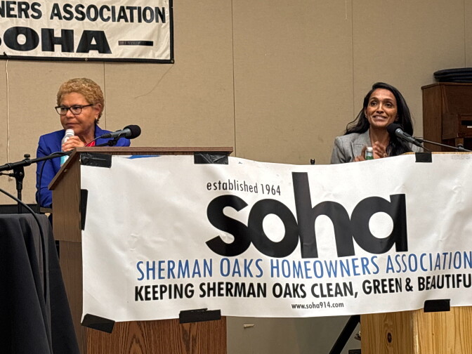 A woman with brown skin and short, light brown hair is behind a lectern. She is wearing a bright blue suit and wearing glasses. To the right of the image, a woman with brown skin and long dark brown hair is behind a separate lectern and microphone. She is wearing a light gray suit. 