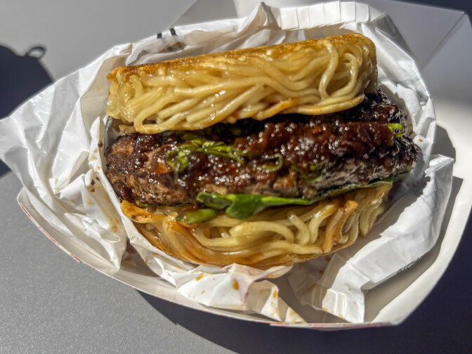 A close-up of a ramen burger — a beef patty glazed in dark sauce with arugula, sandwiched between two seared noodle buns — resting in white food paper.