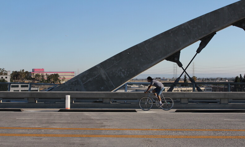 A person rides a bike in a bike lane on a road in front of a large concrete arch of a new bridge.