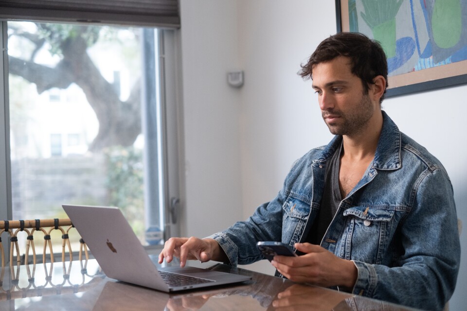 A man with light-tone skin works on a laptop at a table, he also has a phone in his hand. He wears a jean jacket.