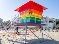A freshly painted lifeguard tower has stripes, from bottom to top, in blue, green, yello, orange and red. It sits on the sand in front of condo buildings.