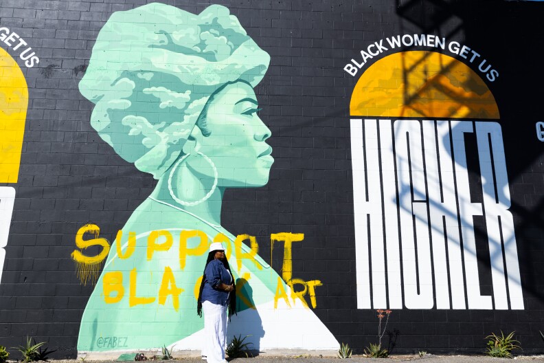 a woman stands in front of a mural that depicts a black woman in a headwrap with the words "support black art" below